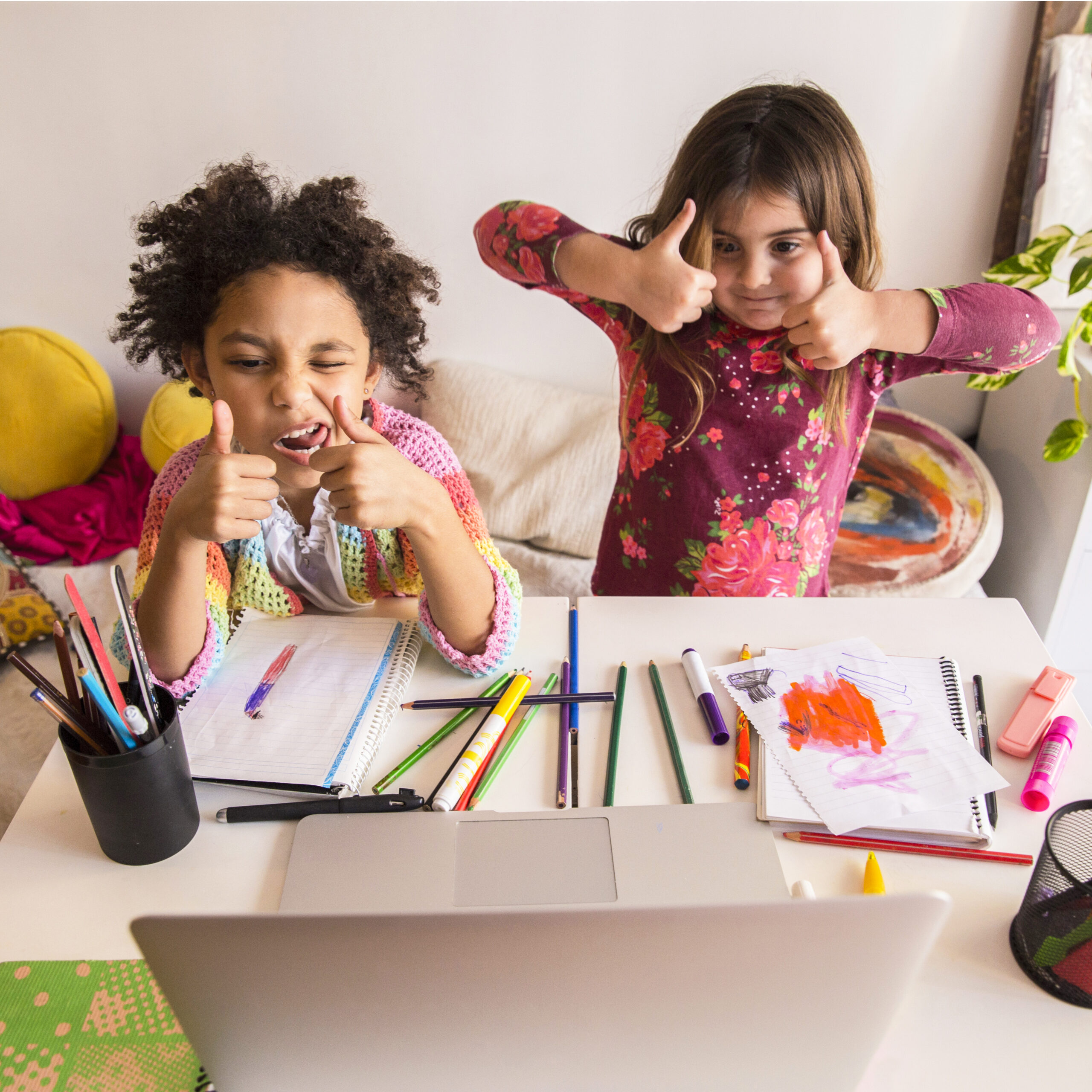 Two young girls enthusiastically show thumbs up while surrounded by colorful art supplies, reflecting a fun and imaginative activity at home.